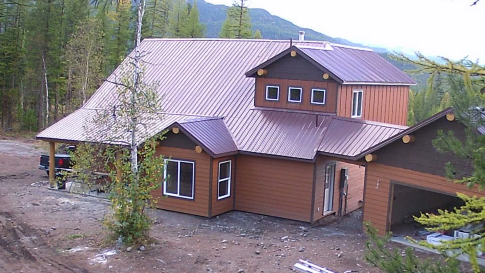 The same house finished from the same spot: brown lap siding, a red standing seam metal roof and a covered entry on log posts