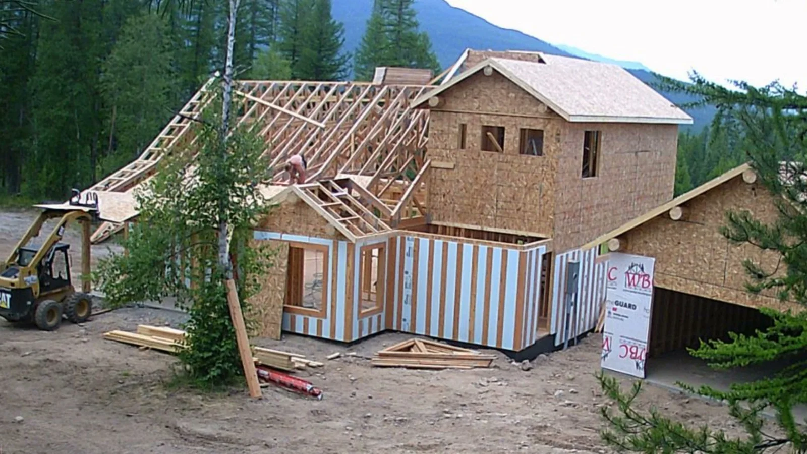 The Glacier Park Retreat at the framing stage: roof trusses going up over sheathed walls, house wrap and a skid steer in the drive