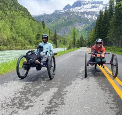 Two adaptive handcycle riders on a mountain road beside a river