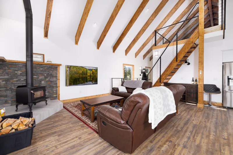 The great room of a custom timber-frame home above Kalispell: exposed beams under a vaulted ceiling, a stone hearth and cable-rail stairs to the loft